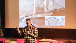 Man föreläser. I bakgrunden en bro som blivit bombad.